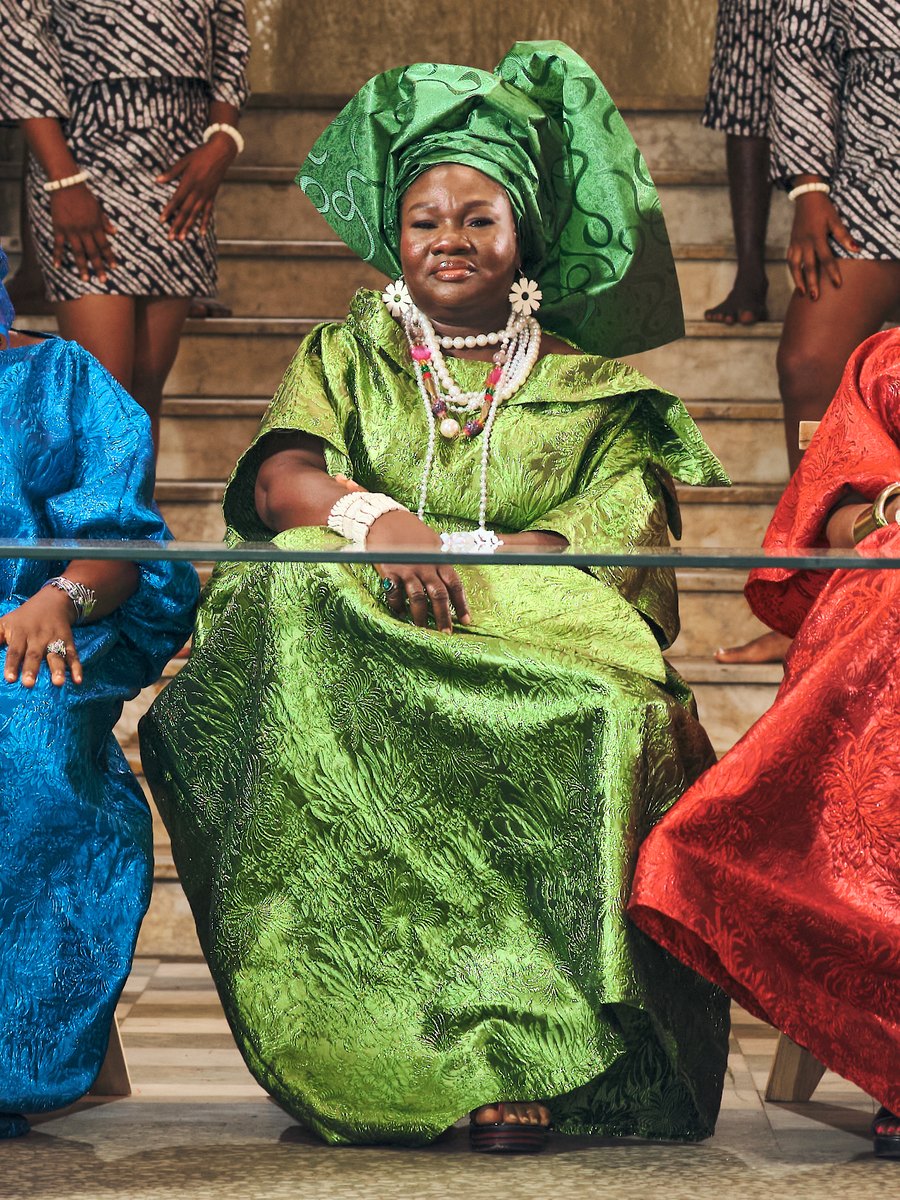 A keeper in a green gele and beads, seated at the table