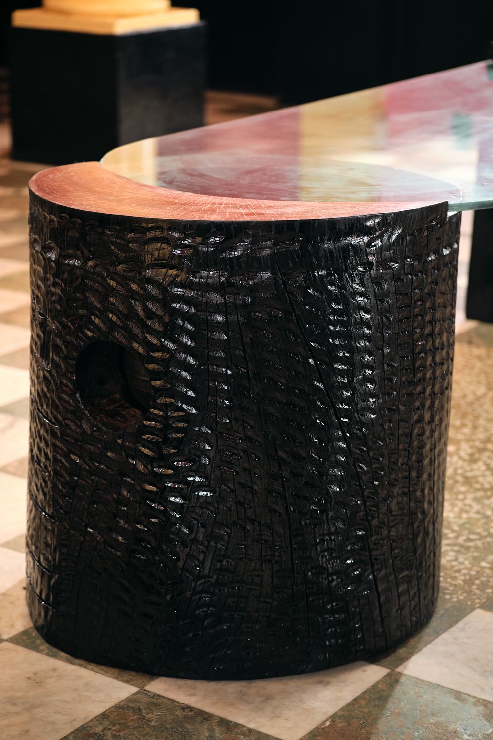 The round base, carved in a scale-like texture, with its reddish wood top beneath the glass
